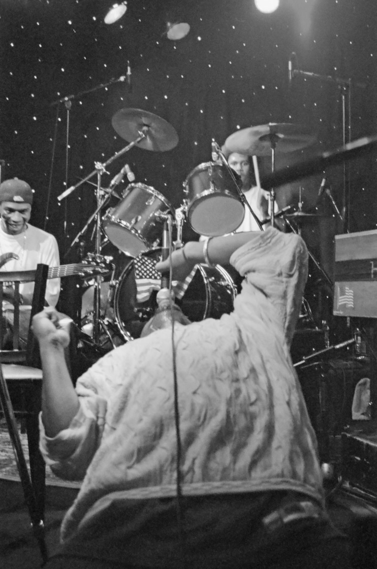 Black and white view from behind the pedal steel player as he flings an arm up, with the drummer and a smiling guitarist playing beyond him.